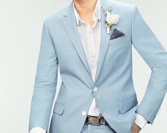 May include: A light blue suit with a white dress shirt and a small white flower on the lapel. The suit has a two-button closure and a matching pocket square. A brown belt is visible.
