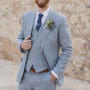 May include: A light blue three-piece suit featuring a textured blazer, a matching vest with five buttons, and trousers. The suit is paired with a crisp white collared shirt, a solid navy blue necktie with a silver tie bar, and a floral boutonniere.
