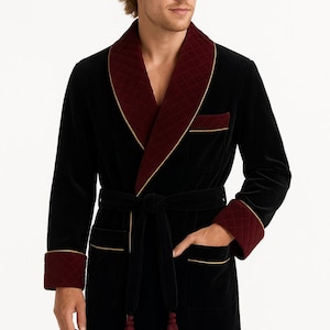 Op de afbeelding: Zwarte fluwelen badjas met een gewatteerde bordeauxrode kraag, manchetten en zakafwerking. De badjas heeft een bijpassende riem met kwastjes en gouden biezen. Een luxe kledingstuk.