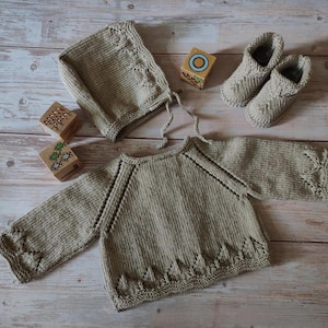 May include: A beige knitted baby set comprising a sweater, bonnet, and booties. The sweater features decorative edging, and the bonnet has ties. Wooden blocks with painted designs are also included in the image.