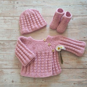 Peut inclure: Ensemble de vêtements pour bébé rose clair tricotés à la main, comprenant un chapeau, un cardigan et des chaussons. Le cardigan a des boutons en bois et un motif texturé. Un petit bouquet de marguerites blanches accompagne l'ensemble.
