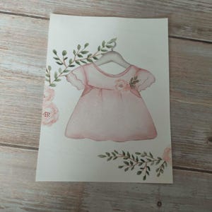 Può includere: Illustrazione ad acquerello di un vestitino rosa per neonati su una gruccia, ornato da elementi floreali e fogliame. L'opera presenta tenui tonalità di rosa e dettagli delicati, evocando tenerezza. L'abito è il punto focale.