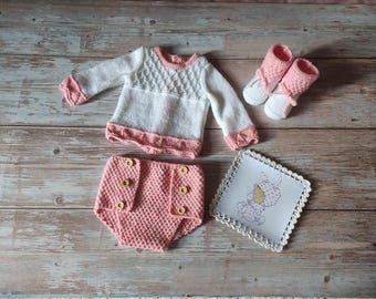 May include: A knitted baby outfit in pink and white. The outfit includes a sweater with a diamond pattern, a diaper cover with buttons, and a pair of booties.