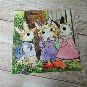 May include: A watercolour illustration of three rabbit characters in a garden scene. One rabbit wears a blue backpack, another a purple dress, and the third a pink dress. Two red apples are in the foreground.