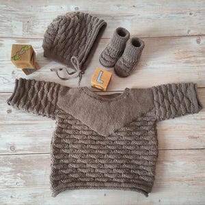 Puede incluir: Un jersey de punto marrón para bebé con un patrón de punto de cable. El jersey tiene un cuello puntiagudo y mangas largas. El jersey está colocado sobre una superficie de madera con un gorro de punto marrón y un par de botines de punto marrón.