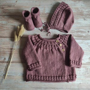 Op de afbeelding: Een gebreide baby-outfit in een stoffige roze kleur. De set bevat een trui met een decoratieve halslijn, een bijpassende muts en slofjes.