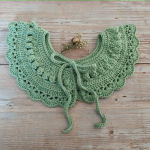 May include: A sage green crocheted collar with a scalloped edge and tie closure. The collar has a detailed, textured pattern and is displayed on a light wood surface. A small dried flower sprig rests in the centre.