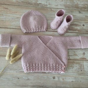 Peut inclure: Ensemble de bébé tricoté lilas comprenant un bonnet, un cardigan croisé et des chaussons. L'ensemble est en fil doux et présente un motif de tricot délicat. Le cardigan a deux boutons. Les articles sont disposés sur une surface en bois.