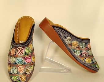 May include: A pair of black embroidered slippers with a colourful swirl pattern. The slippers have a brown leather sole and a wedge heel.