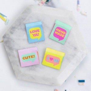 Peut inclure: Quatre post-it colorés avec des motifs de bulles de dialogue. Les notes sont bleues, vertes, roses et jaunes, chacune avec un message différent dans une couleur contrastante : "LOVE YA!", "HAND MADE", "CUTE!" et "I LOVE U!". Les notes sont disposées sur un hexagone en marbre blanc.