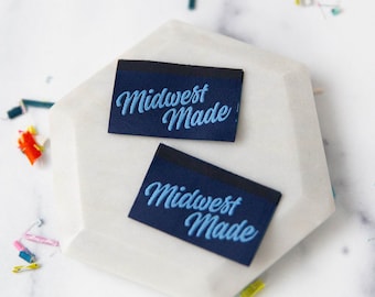 Könnte beinhalten: Zwei marineblaue Stoffetiketten mit dem Text "Midwest Made" in hellblauer Schrift. Die Etiketten sind rechteckig mit einem schwarzen Rand oben. Sie liegen auf einer weißen, geometrischen Oberfläche.