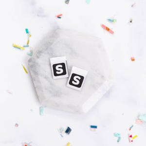 Puede incluir: Dos pequeñas etiquetas blancas con un cuadrado negro y una "S" blanca se muestran sobre una superficie de mármol blanco. Las etiquetas son rectangulares y tienen un diseño limpio y minimalista. Confeti colorido está esparcido alrededor.