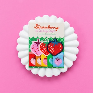 May include: A set of Strawberry by Kimberly Kight labels featuring 5 sew-in and 3 iron-on labels. The packaging displays three large strawberry patches in pink, red, and dark blue, with five smaller colorful fabric labels below featuring strawberry icons.