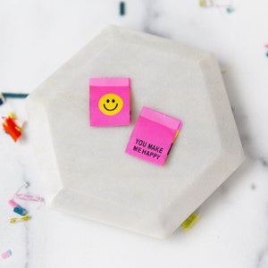 May include: Two pink fabric labels with text and graphics rest on a white, geometric surface. One label features a yellow smiley face, and the other reads "YOU MAKE ME HAPPY" in white text. The labels are rectangular with folded edges.