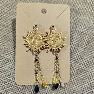 Puede incluir: Pendientes dorados con un diseño celestial de sol y luna. Los pendientes tienen cadenas colgantes con cuentas negras, amarillas y blancas. Los pendientes se muestran en una tarjeta marrón.