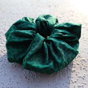 May include: A dark green fabric hair scrunchie featuring a repeating pattern of small, dark, geometric leaf-like shapes. The accessory has a gathered, voluminous texture with soft folds throughout the circular design, creating a full appearance.