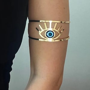 May include: Gold-toned arm cuff featuring an eye design with a blue center. The cuff has a stylized sunburst pattern above the eye and is worn on the upper arm. The design is a unique piece of jewelry.