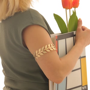 May include: A gold-colored arm cuff with a leaf design. The cuff is worn on the upper arm. The person is wearing a green shirt and holding a book and flowers.