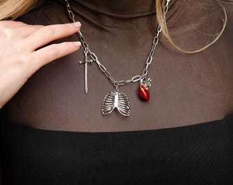 Puede incluir: Un collar de cadena plateada con un dije de espada, caja torácica y corazón rojo. El collar lo lleva una persona con el pelo largo y rubio.