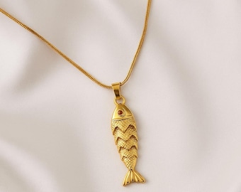 May include: Gold-colored fish pendant necklace. The pendant features a detailed fish design with textured scales and a small red accent for the eye. The necklace is displayed on a white background.