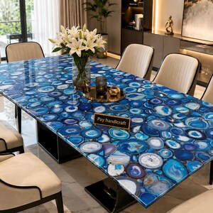 Puede incluir: Una mesa de comedor rectangular con una superficie pulida compuesta por numerosas rodajas de ágata azul en varios tonos de azul, cobalto y blanco. Una pequeña placa negra con el texto dorado Pay handicraft se encuentra sobre la mesa.