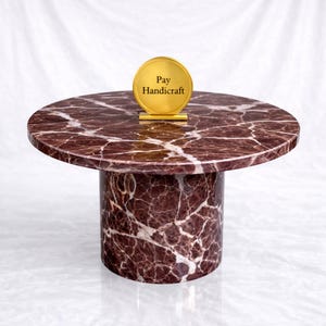 May include: A round, marble-topped table with a cylindrical base. The tabletop is a deep burgundy with white veining. A gold-colored sign with the text "Pay Handicraft" stands on the table.