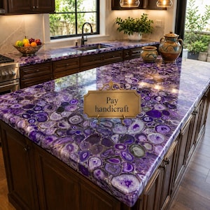 May include: A kitchen island countertop made of polished purple agate stone slabs with a natural geode pattern. A rectangular gold-toned sign with an ornate frame sits on the surface, featuring the black text Pay handicraft in the center.