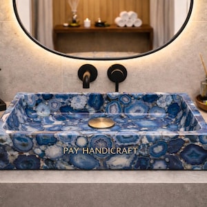 Könnte beinhalten: Rechteckiges Waschbecken aus blauem Achatstein mit goldenem Abfluss. Das Waschbecken steht auf einer grauen Arbeitsplatte, mit schwarzen Armaturen und einem runden Spiegel im Hintergrund. Das Waschbecken hat ein einzigartiges, natürliches Muster.