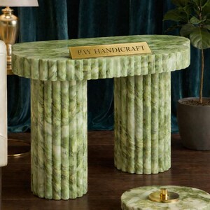 Könnte beinhalten: Eine grüne Marmor-Konsole mit geriffeltem Design. Der Tisch hat zwei zylindrische Beine und eine ovale Platte. Ein goldenes Schild auf dem Tisch lautet "PAY HANDICRAFT". Der Tisch ist ein dekoratives Möbelstück.