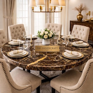 Op de afbeelding: Een ronde eettafel met een bruin marmeren blad en gouden accenten, gedekt voor een maaltijd. De tafel is omringd door beige gestoffeerde stoelen. Een witte vaas met bloemen staat in het midden en de tafelsetting omvat goudgerande borden en bestek. Het bord op de tafel luidt "PAY HANDICRAFT".