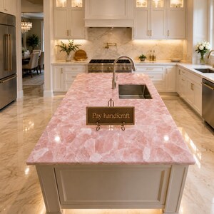 May include: A kitchen island featuring a polished pink rose quartz countertop with a rectangular sink and a metal faucet. A small, rectangular dark brown sign with gold lettering that reads "Pay handicraft" sits on a metal stand on the counter.