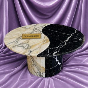 Könnte beinhalten: Einzigartiger Marmor-Couchtisch mit Yin-Yang-Design. Eine Hälfte ist weiß und gold, die andere schwarz mit weißen Adern. Der Tisch hat einen rechteckigen Sockel und eine ovale Platte. Der Text "PAY HANDICRAFTS" ist zu sehen.
