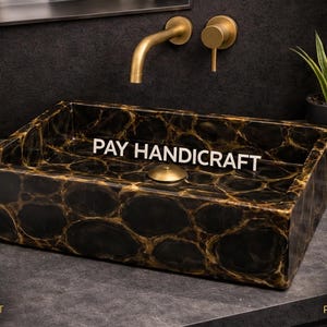 Könnte beinhalten: Rechteckiges Badezimmerwaschbecken aus schwarzem und goldenem Stein mit dem Text "PAY HANDICRAFT". Das Waschbecken hat einen goldenen Abfluss und steht vor einer dunklen Wand mit einem goldenen Wasserhahn. Eine Kerze und eine Pflanze sind sichtbar.