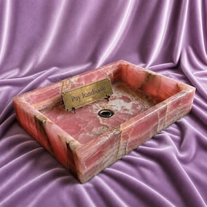 Könnte beinhalten: Ein rechteckiges Waschbecken aus rosa Onyx mit Abfluss und einem kleinen Messingschild mit der Aufschrift "Pay handicraft". Das Waschbecken hat eine polierte Oberfläche und steht vor einem violetten Samthintergrund. Ein einzigartiges Badezimmer-Element.