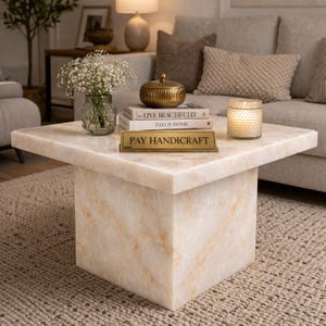 Puede incluir: Mesa de centro cuadrada con aspecto de m&aacute;rmol en crema y dorado. Presenta un patr&oacute;n veteado y sostiene un jarr&oacute;n de cristal con flores blancas, libros, un recipiente dorado, una vela y un letrero de lat&oacute;n con "PAY HANDICRAFT".