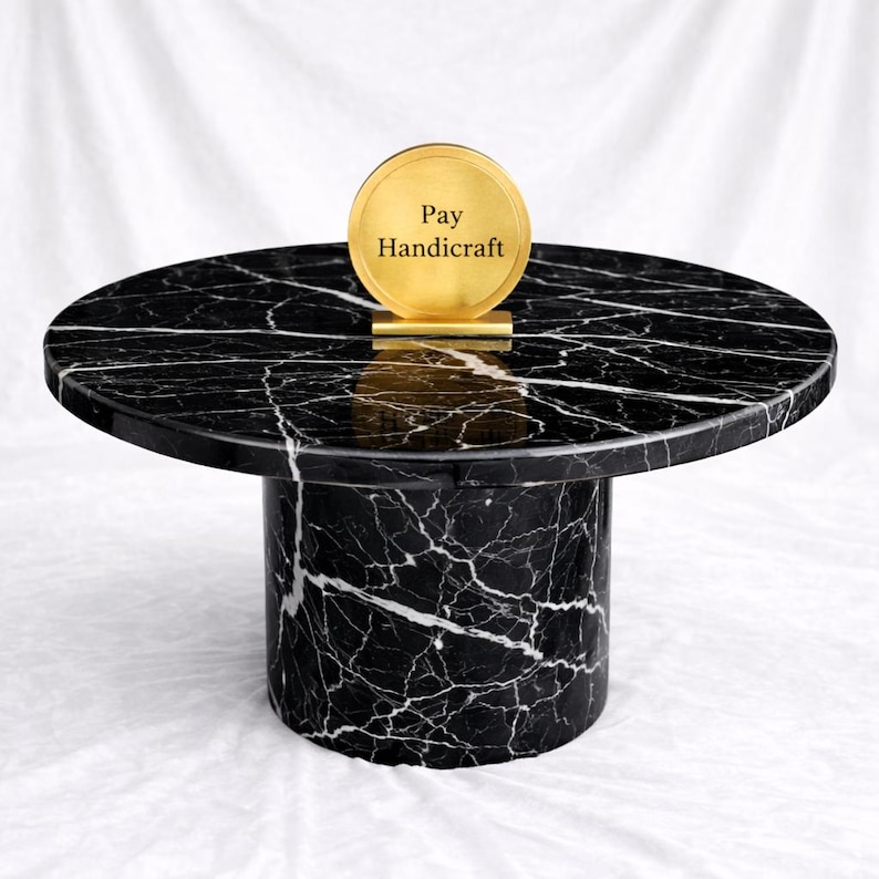 May include: A black marble side table with white veining. The table has a round top and a cylindrical base. A gold-colored disc with the text "Pay Handicraft" is on top of the table.