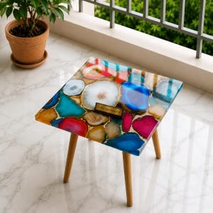 May include: A square side table featuring a tabletop composed of multicolored agate slices embedded in clear resin. The table has four light-colored wooden legs. A small rectangular plaque on the surface displays the text Pay handicraft.