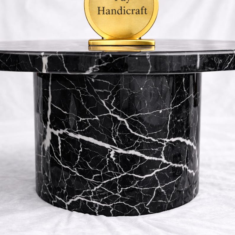 May include: A black and white marble table with a cylindrical base. The tabletop is round and the base is a solid cylinder. The marble has a striking white veining pattern. A gold-colored emblem with the text "Pay Handicraft" is on top.