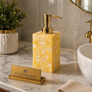May include: A yellow and white mosaic soap dispenser with a gold pump. The dispenser is rectangular and sits on a marble countertop. A gold sign that reads "PAY HANDICRAFT" is in front of the dispenser. A small plant in a white pot is to the left.