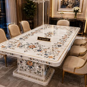 Op de afbeelding: Een rechthoekige eettafel van wit marmer met een centrale voet, voorzien van ingelegde bloemmotieven in oranje, blauw en groen. De tafel is omringd door acht beige gestoffeerde stoelen met gouden metalen frames. Een bordje vermeldt Pay handicraft.