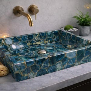 Könnte beinhalten: Rechteckiges Waschbecken aus blauem und goldgeädertem Stein mit dem Text "PAY HANDICRAFT". Das Waschbecken ist auf einer Marmorarbeitsplatte montiert, mit einem goldenen Wasserhahn und einer kleinen Pflanze im Hintergrund. Eine Kerze und Seife sind ebenfalls sichtbar.