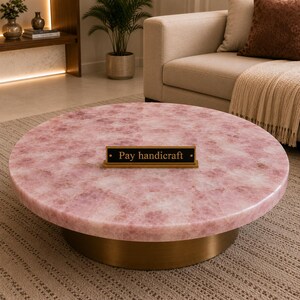 May include: A round coffee table featuring a polished pink rose quartz stone top with a natural crystalline texture and a solid cylindrical brass base. A small rectangular black sign with gold text reading Pay handicraft sits on top.