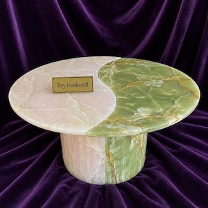 Könnte beinhalten: Ein handgefertigter, ovaler Couchtisch mit Yin-Yang-Design. Der Tisch besteht aus Marmor, mit einer rosa-weißen Seite und einer grün-goldenen Seite. Der Sockel ist zylindrisch und passt zum Farbschema. Ein kleines Messingschild trägt die Aufschrift "Pay handicraft."