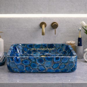 Könnte beinhalten: Ein rechteckiges Waschbecken aus blauem Achatstein mit einem einzigartigen Muster. Das Waschbecken ist der Mittelpunkt eines Badezimmers, ergänzt durch goldene Armaturen, eine weiße Vase mit Tulpen und einen weißen Wecker. Das Waschbecken trägt den Text "PAY HANDICRAFT".