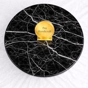 May include: A round, black marble tray with white veining. A gold-colored, circular sign with the text "Pay Handicraft" is centered on the tray. The tray is on a white surface.