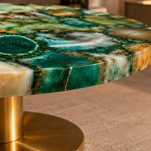 May include: A round coffee table featuring a polished surface made of green and amber agate stone slices with natural crystalline patterns. The tabletop is supported by a cylindrical pedestal base with a smooth, brushed metallic gold finish.