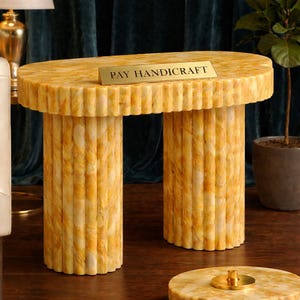 Könnte beinhalten: Ein gelb-weißer Tisch in Marmoroptik mit zwei zylindrischen Beinen. Die ovale Tischplatte hat einen geriffelten Rand. Ein kleines goldenes Schild oben zeigt "PAY HANDICRAFT". Der Tisch steht auf einem dunklen Holzboden.
