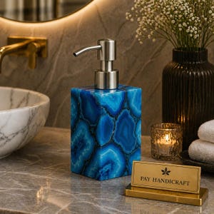 Puede incluir: Un dispensador de jabón azul y blanco con un patrón de ágata y un cabezal de bomba plateado. El dispensador está sobre una encimera de mármol. Un letrero dorado dice "PAY HANDICRAFT". Una vela encendida y un jarrón de flores están en el fondo.