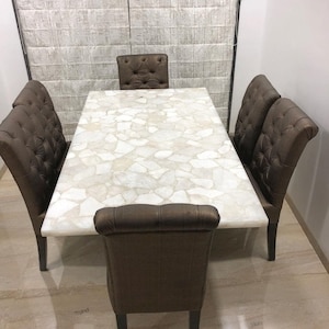 May include: A white dining table with a unique, agate-like surface. The table is surrounded by six brown upholstered chairs with button tufting.