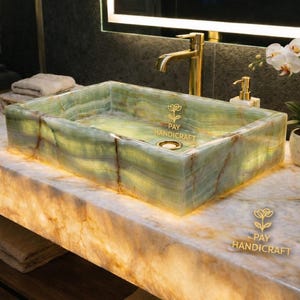 Könnte beinhalten: Ein rechteckiges, hinterleuchtetes Waschbecken aus grünem Onyx mit einer goldenen Armatur. Das Waschbecken steht auf einer Marmorplatte, mit einer brennenden Kerze und einer Orchideenpflanze. Das Waschbecken trägt den Text "PAY HANDICRAFT".
